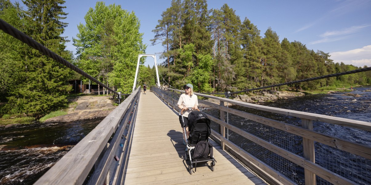 Kävelysillalla nainen lastenrattaiden kanssa kävelee katsojaa kohti. Sillan alla vettä, taustalla puita.