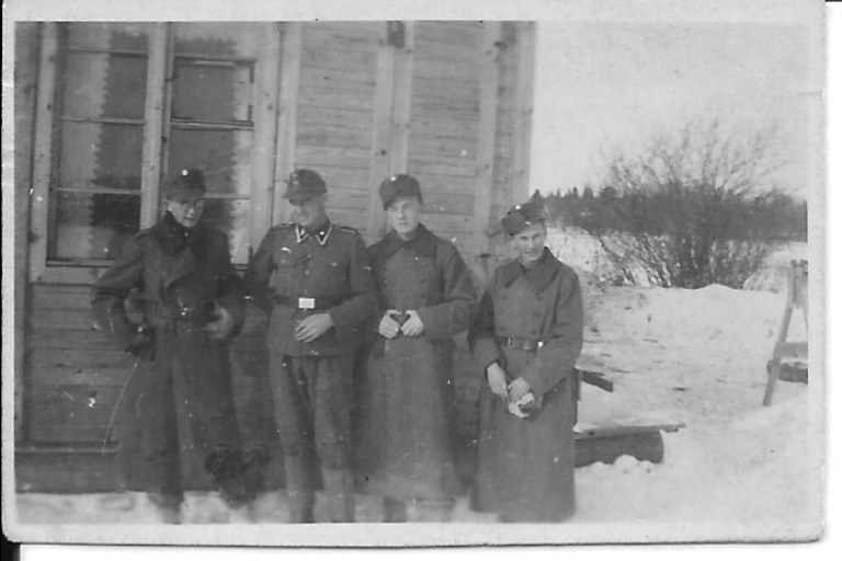 Neljä sotilasta seisoo talvella puutalon edustalla.
