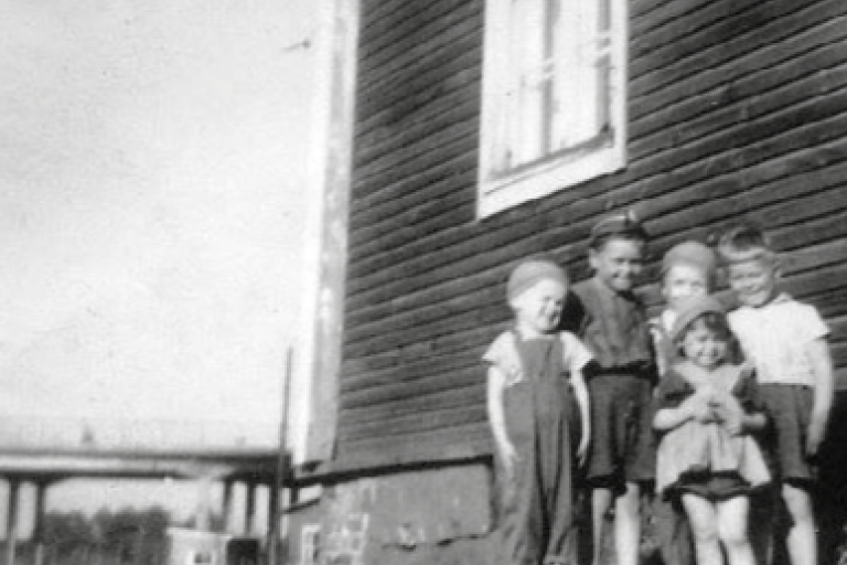 Mustavalkoisessa kuvassa lapsia seisoo puutalon edustalla kesäpäivänä.