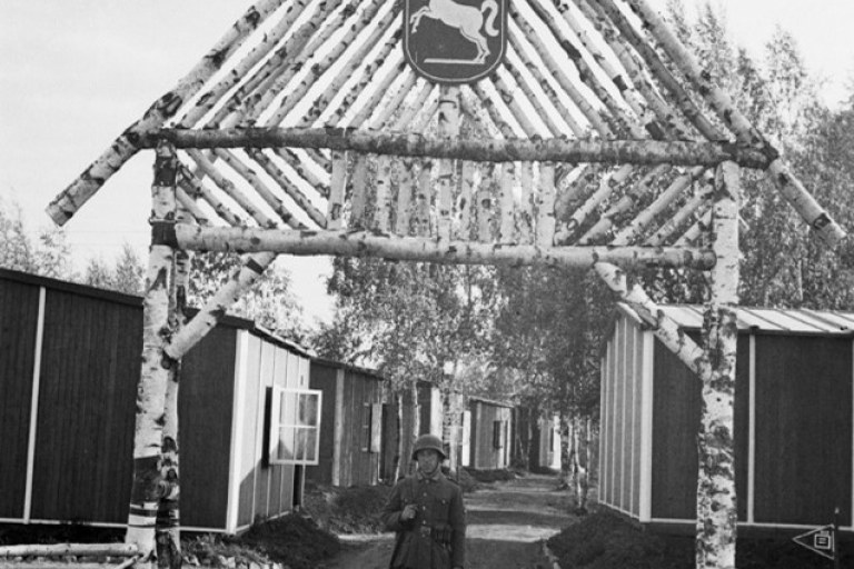 Mustavalkokuvassa sotilas seisoo puisen portin edustalla.