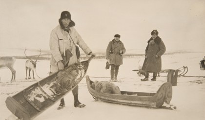 Vanha, mustavalkoinen valokuva, jossa on kolme talviasuista henkilöä, joista yksi esittelee ahkiota, johon on pakattu tavaraa.