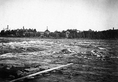 Vanha, mustavalkoinen valokuva jossa on leveä joki, vastarannalla näkyy rakennuksia.