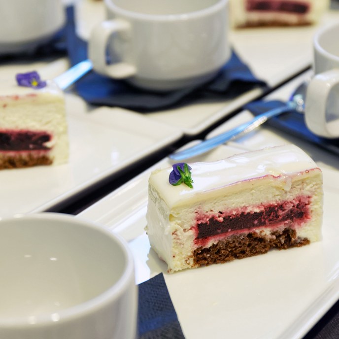 A slice of cake on a plate, with coffee cups in the background
