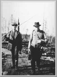 Vanha, mustavalkoinen valokuva, jossa on kaksi miestä metsäaukiolla.