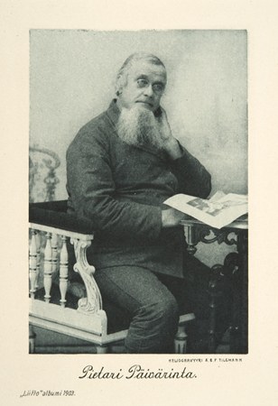 Mustavalkoinen, vanha valokuva jossa parrakas mies istuu koristeellisella tuolilla ja lukee pöydän ääressä lehteä.